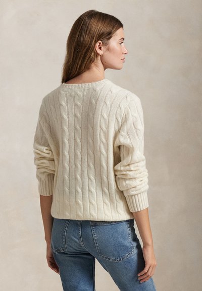 Camisola de malha creme com padrão de trança, punhos canelados e decote redondo, combinada com jeans de ganga azul.