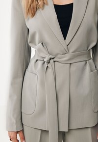 Blazer grigio con cintura, grandi tasche frontali, texture liscia e design strutturato. Presenta una rever a punte e dettagli con cinturino in vita.