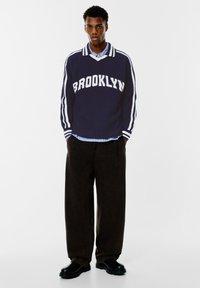 Námořnický pletený svetr s nápisem "BROOKLYN" v bílé barvě, spárovaný se světle modrou košilí s límečkem. Celkový vzhled doplňují tmavé široké kalhoty a černé boty.