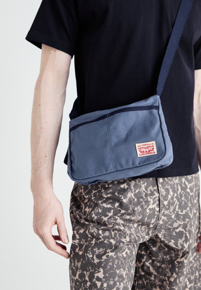 Mala mensageiro de lona azul com um rótulo vermelho; possui alça ajustável e costura contrastante. Material texturizado, forma retangular prática.