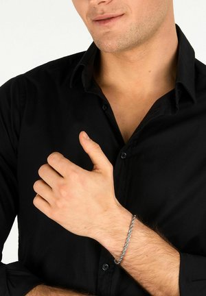 Uomo che indossa una camicia nera con bottoni che mostra un braccialetto a catena argentato intrecciato al polso sinistro su sfondo bianco.
