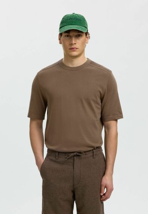 Młody mężczyzna w brązowej koszuli z krótkim rękawem, brązowych spodniach ze sznurkiem oraz zielonej czapce z napisem „LA PISCINE”, stojący na neutralnym tle.