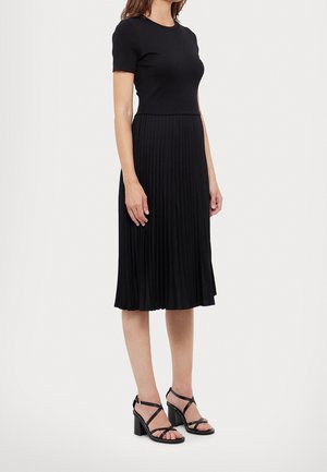 Femme portant un haut noir à manches courtes, une jupe midi plissée et des sandales noires à talons blocs à brides, sur un fond uni.