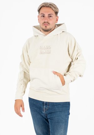 Kremowa bluza z kapturem z przednią kieszenią, z delikatnym logo na środku. Gładka tekstura i luźny krój, w połączeniu z niebieskimi dżinsami.