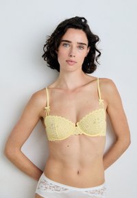 Gelbes Spitzen-BH mit gewellten Kanten, der zarte Schleifenakzente an den Trägern und ein florales Ausstanzmuster aufweist.