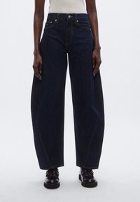 Laiade tumedad teksapüksid kontrastsete õmblustega, kõrge vöökoht ja viie taskuga disain. Vöö juures istuvad tihedalt, jalgadest laiad.