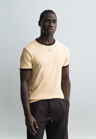 Homme à la peau foncée portant un t-shirt jaune pâle avec des bordures noires et un pantalon noir, debout devant un fond gris clair uni.