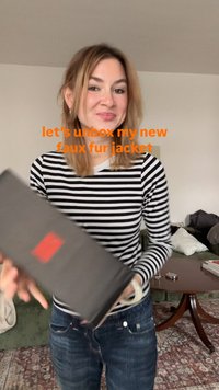 Unboxing di una giacca in pelliccia sintetica. La modella indossa un top a maniche lunghe a righe bianche e nere. Il pacco è nero, con un logo rosso. Sono visibili fibre naturali.