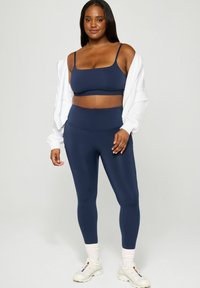 Navy blue athletic set featuring a fitted crop top and high-waisted leggings, made from smooth, stretchy material. White zip-up hoodie and shoes.