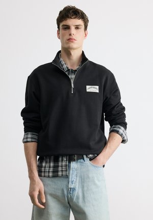 Fiatal férfi fekete negyed-cipzáras pulóvert visel fekete-fehér kockás ing fölött és világoskék farmernadrágot, áll egy kézzel a zsebében.