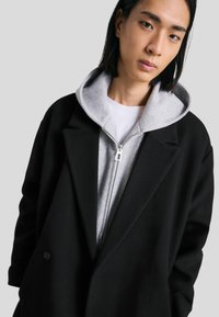 Zwarte oversized jas met revers over een grijze rits-hoodie. De hoodie heeft een zachte textuur en een capuchon, gelaagd over een wit T-shirt.