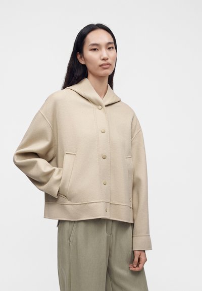 Femme aux longs cheveux noirs portant une veste à capuche beige boutonnée et un pantalon gris clair, debout devant un fond blanc uni.