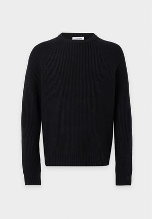 Jeune homme debout les mains dans les poches, portant un pull côtelé noir, un pantalon beige et des mocassins noirs sur un fond clair uni.
