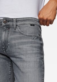 Jeans de mezclilla gris con corte recto, sutiles arrugas y una etiqueta de marca negra en la cintura. Incluye detalle de botón de metal y cremallera.