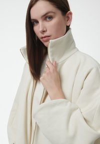 Chaqueta de forro polar blanca con alto cuello y cremallera dorada, que presenta mangas de gran tamaño y una textura suave, parcialmente abierta en la parte delantera.