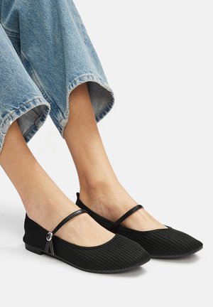 Schwarze gerippte Ballerinas mit einem dünnen Riemen und silbernem Hardware-Detail. Weicher Stoff, flaches Profil, kombiniert mit blauen, weiten Denim-Hosen.