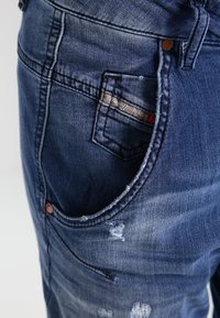 Mörkblå denimjeans med en sliten design. Har en framficka, kontrasterande sömmar och metallknappdetaljer.