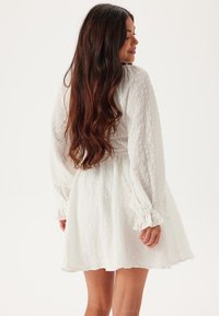 Vit texturerad klänning med långa ärmar och volangärmar, med en figurnära bodice och en utflädd kjol. Subtilt mönster, lättviktigt tyg.