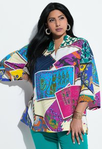 Farbenfrohe, übergroße Bluse in verschiedenen Druckblöcken mit Tier- und Blumendesigns, mit Kragen und lockeren Ärmeln. Kombiniert mit türkisfarbenen Hosen.