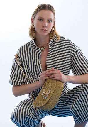 Beige strukturierte Tasche mit geschwungener Form, goldener Kettenriemen und dekorativem Akzent. Modell in gestreiftem blau-weißem Outfit, in hockender Pose.