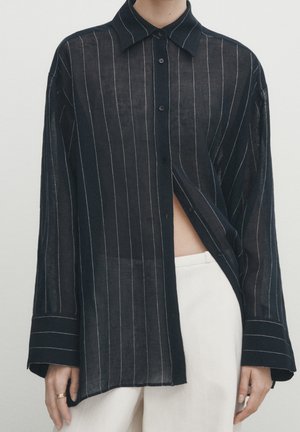 Osoba ubrana w czarną przezroczystą koszulę z długim rękawem z białymi pionowymi paskami, częściowo rozpiętą, w zestawie z białymi spodniami z wysokim stanem.