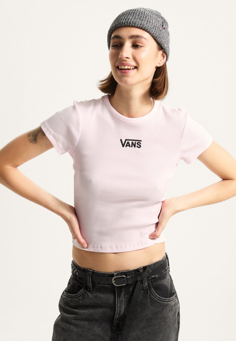 Helles rosa, geripptes Crop-Top mit rundem Ausschnitt und schwarzem "VANS"-Logo. Kombiniert mit hochgeschnittenen dunklen Jeans und einer grauen Strickmütze.