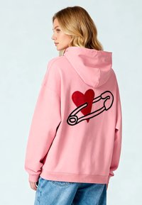 Felpa oversize rosa in cotone con un ricamo a forma di cuore rosso e un design di spilla di sicurezza sulla schiena. Tasca a marsupio e cappuccio con cordino.