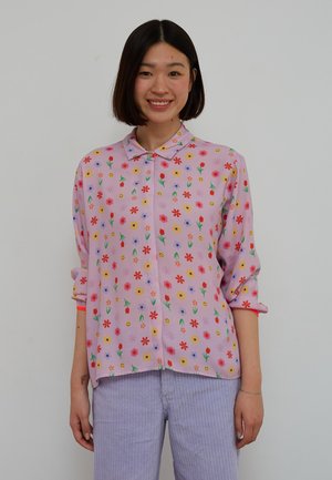 Young woman with short dark hair wearing a pastel purple button-up shirt with colorful flower and smiley face patterns and light purple pants.