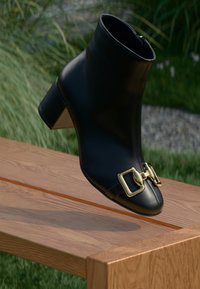 Bottine en cuir noir avec détail de boucle en or, bout pointu et talon carré, présentée sur un fond naturel sur une surface en bois.