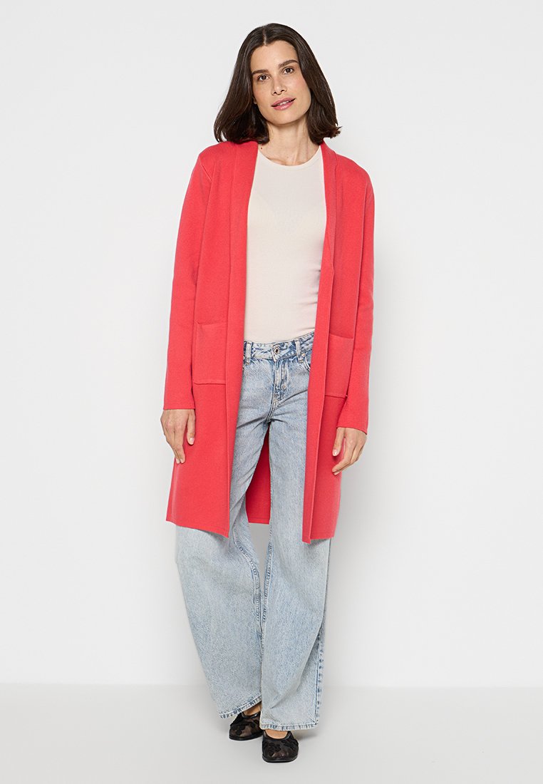 Frau in einem korallenfarbenen langen Cardigan, weißem Oberteil, hellblauen weit geschnittenen Jeans und schwarzen Schuhen, die vor einem weißen Hintergrund steht.