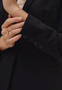 Zwarte getailleerde blazer met een gladde textuur, voorzien van vier zwarte knopen op de manchetten en een snit tot onder de elleboog. Ring met twee bolvormige accenten.