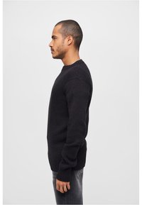 Suéter negro de punto con puños y dobladillo acanalados, que presenta un cuello redondo y costuras de hombro estructuradas. Se lleva con unos vaqueros gris ajustados.