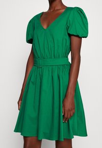 Grön bomullsklänning med V-ringning, puffärmar, åtdragen midja och full kjol. Har ett bälte med en rektangulär spänne.