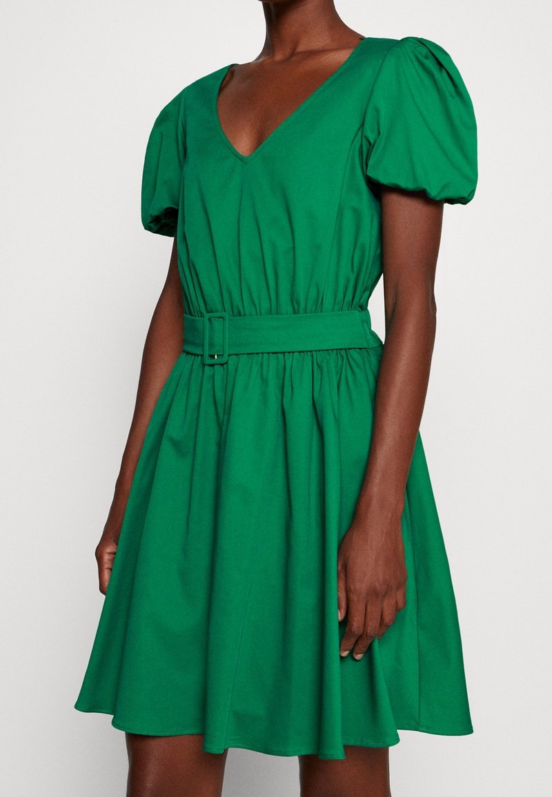 Grön bomullsklänning med V-ringning, puffärmar, åtdragen midja och full kjol. Har ett bälte med en rektangulär spänne.