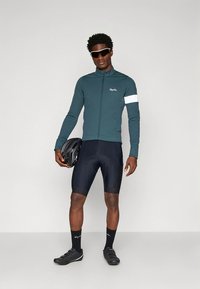 Donker tealkleurig fietsshirt met witte accenten, gecombineerd met zwarte gevoerde shorts en zwarte fietsschoenen. Model houdt een zwart helm vast.