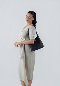 Crna kožna torba s zlatnim detaljima, glatke teksture, u trapezoidnom obliku. Model nosi haljinu svijetlih tonova s drapiranim detaljima.