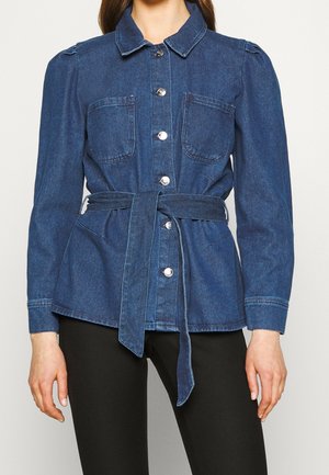 Femme portant une veste en jean bleue à boutons, avec un col, des poches poitrine, des manches longues et une ceinture assortie nouée à la taille.