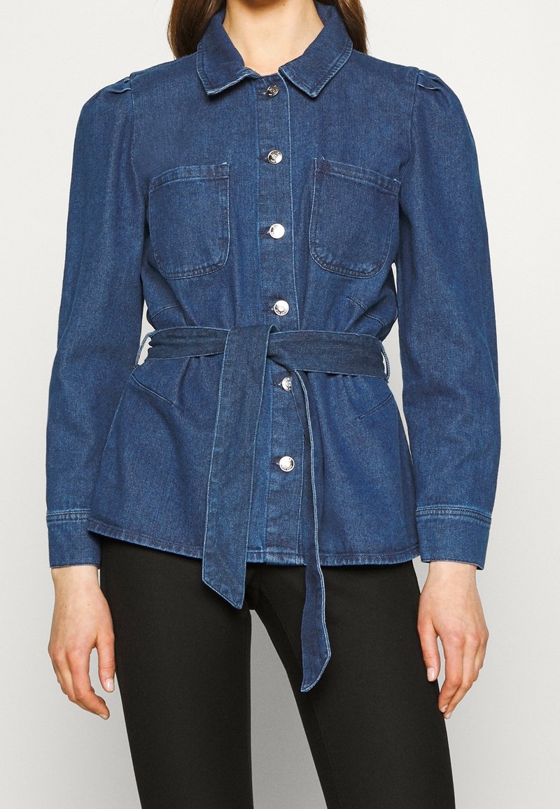 Femme portant une veste en jean bleue à boutons, avec un col, des poches poitrine, des manches longues et une ceinture assortie nouée à la taille.