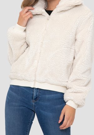 Femme portant une veste polaire sherpa blanche zippée avec poignets et ourlet côtelés, associée à un jean en denim bleu, main près du col.