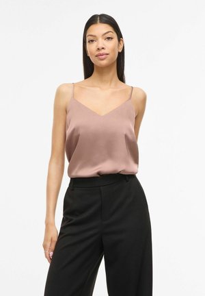 Kvinde med langt sort hår iført en støvet lilla ærmeløs bluse og sorte bukser med høj talje, stående foran en ensartet hvid baggrund.