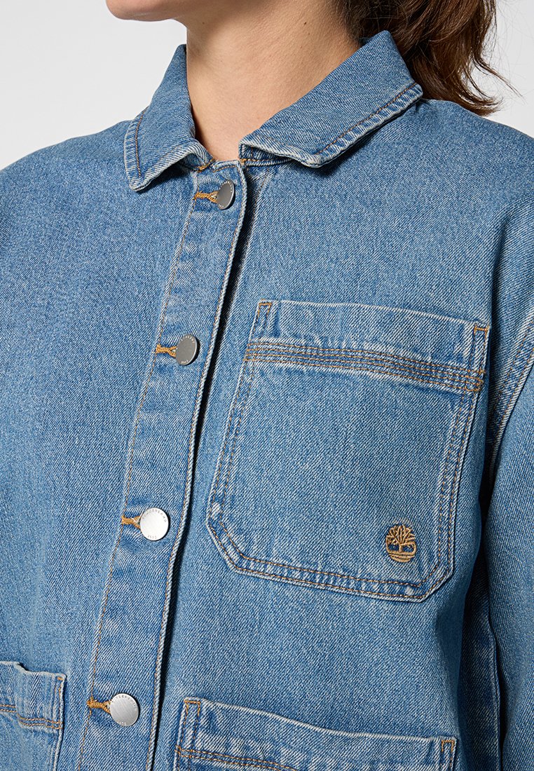 Donna che indossa una giacca di jean azzurro chiaro con bottoni a pressione in metallo e una tasca sul petto con un piccolo logo ad albero.