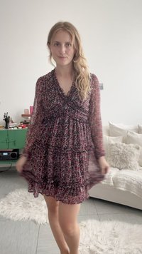 Νεαρή γυναίκα με μακριά ξανθά μαλλιά που φοράει σκούρο φόρεμα με μακριά μανίκια και λουλούδια, στέκεται σε φωτεινό σαλόνι με άσπρο καναπέ και πράσινη ντουλάπα πίσω της.