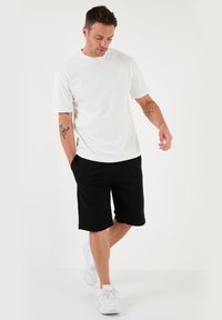 T-shirt bianca a maniche corte abbinata a pantaloni corti neri fino al ginocchio, con un design semplice. Il modello indossa sneakers bianche.