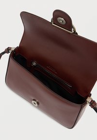 Δερμάτινη τσάντα ώμου σε σκούρο καφέ με επάνω καπάκι, ασημένιο μεταλλικό εξάρτημα, εσωτερική τσέπη με φερμουάρ και ομαλή υφή. Το εσωτερικό είναι μαύρο.