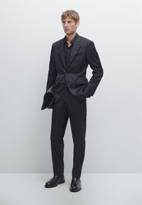 Schwarzer maßgeschneiderter Anzug mit schlanker Passform, einreihigem Sakko, flachfrontiger Hose und schwarzen Lederstiefeln. Glatter Stoff und strukturierter Schnitt.