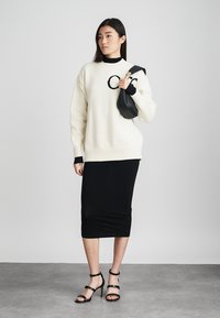 Pull en tricot crème avec des accents noirs, associé à une jupe noire ajustée. La mannequin tient un petit sac noir et porte des talons noirs à brides.