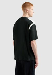 Giovane con i capelli rossi corti che indossa una maglietta nera con dettagli bianchi sulle spalle, in piedi di profilo contro uno sfondo chiaro.