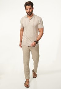 Beige Polohemd mit kurzen Ärmeln, das vertikale Strickmuster aufweist, kombiniert mit locker sitzenden beigen Leinenhosen und braunen Sandalen.