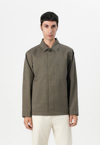 Brown and black checked jacket with a buttoned collar, long sleeves, and a structured fit, paired with light-colored pants.