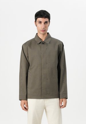 Brown and black checked jacket with a buttoned collar, long sleeves, and a structured fit, paired with light-colored pants.